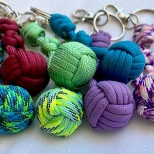 May include: A selection of colourful paracord keyrings. The keyrings display a variety of colours, such as teal, green, purple, and red. Each keyring has a decorative knot design and a silver-coloured metal clasp.