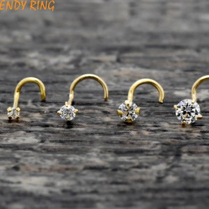 May include: Four gold nose rings with clear, round gemstones of varying sizes. The curved posts are visible, and the jewelry is set against a textured, dark wood surface. The brand name "TRENDY RING" is visible in the top left corner.