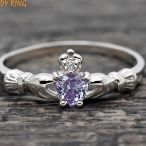 May include: A silver Claddagh ring with a purple heart-shaped gemstone and a smaller clear gemstone in the crown. The ring is a traditional Irish symbol of love, loyalty, and friendship.
