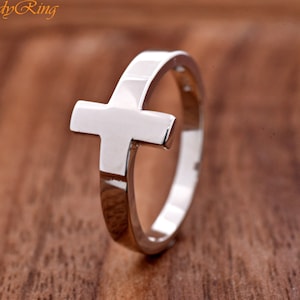 May include: A polished silver-toned ring featuring a prominent cross design integrated into the band. The metal surface is smooth and reflective, showcasing a clean, minimalist aesthetic. The text TrendyRing is visible in the upper left corner.