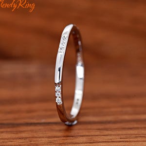 May include: A thin silver-toned metal ring featuring a smooth band with two distinct sections of three small, clear, round-cut stones embedded in the surface. The ring is positioned vertically, displaying a polished finish and reflective surface.