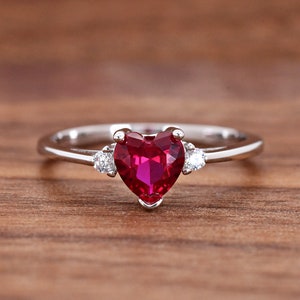 May include: A silver ring with a heart-shaped ruby gemstone and two smaller accent stones. The ring is on a wooden surface.