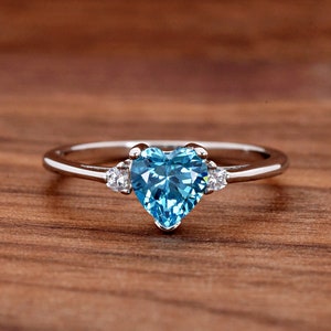 May include: A silver ring with a heart-shaped blue topaz gemstone and two smaller clear gemstones on the band.