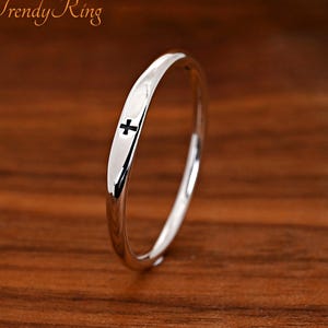 May include: A thin, polished silver-tone ring featuring a small, black cross symbol engraved on the outer surface. The band has a smooth, rounded profile and a reflective metallic finish. The text "Trendy Ring" is visible in the upper left corner.