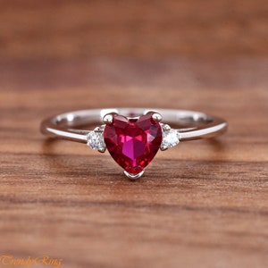 May include: A silver-toned ring featuring a central heart-shaped red gemstone held in a prong setting. The band is accented on each side by a single small, round, clear white gemstone, creating a symmetrical design with a polished metallic finish.