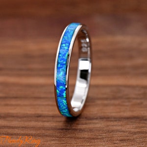 May include: A silver ring with a band of vibrant blue opal. The opal is inlaid in a mosaic pattern, creating a striking contrast against the polished silver. The ring is set against a warm, wooden background.