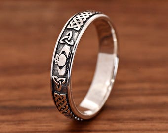 May include: A silver-coloured metal ring featuring an embossed Claddagh symbol at the centre, flanked by repeating triquetra knots and intricate woven patterns along the band. The surface has a darkened, oxidised finish to highlight the detailed engravings.