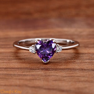 May include: A silver-toned ring featuring a central heart-shaped purple gemstone flanked by two small, clear round stones on either side. The band is smooth and polished, showcasing a clean design that highlights the vibrant violet center stone.
