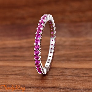May include: A silver eternity band ring featuring a continuous row of small, round, vibrant pink gemstones set in a polished metal channel. The ring is positioned vertically, showcasing the uniform circular arrangement of the pink stones.