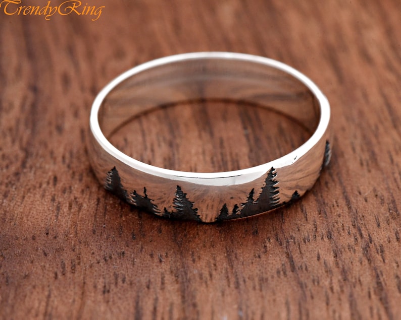 Puede incluir: Un anillo de banda de plata con una silueta de un bosque grabado en la superficie.