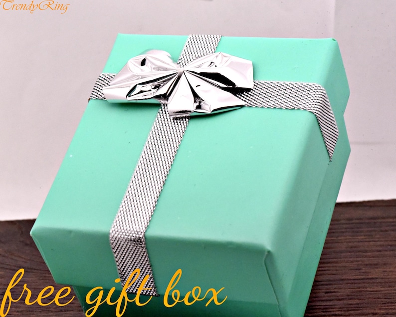 May include: A square turquoise gift box featuring a silver metallic bow and a silver textured ribbon wrapped around the lid. The text TrendyRing is visible in the top left corner, and the text free gift box is written in yellow at the bottom.