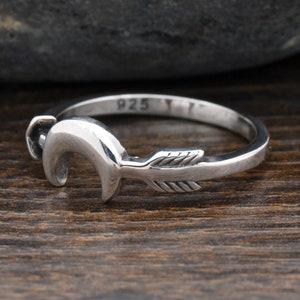 May include: A silver ring with a crescent moon and arrow design. The ring is stamped with the number "925", indicating it is made of sterling silver.