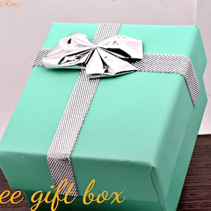 Puede incluir: Una caja de regalo turquesa con un lazo plateado y una cinta plateada con un patr&oacute;n blanco. La caja est&aacute; sobre una superficie de madera. El texto "free gift box" est&aacute; impreso en la parte delantera de la caja.