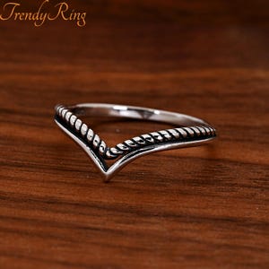 May include: A silver-toned V-shaped ring featuring a twisted rope-like texture along the top band. The metal has darkened recessed areas to highlight the rope detail. The text "Trendy Ring" is visible in the upper left corner of the image.