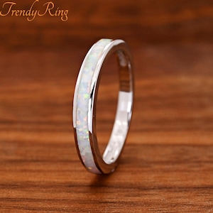 May include: A silver-toned ring featuring a central inlay of iridescent white opal with subtle flashes of green and pink. The band has a polished metallic finish and a smooth, rounded profile. The text Trendy Ring is visible in the top left corner.
