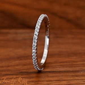 May include: A thin silver-toned band ring featuring a continuous row of small, clear, round-cut stones set along the outer edge. The metal has a polished, reflective finish, and the stones are arranged in a single line to create a sparkling effect.