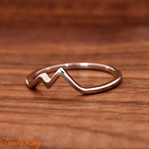 May include: A silver-colored ring with a mountain range design. The ring is set against a wooden background. The ring's design features a series of peaks and valleys, creating a minimalist mountain silhouette. The ring is a piece of jewelry.