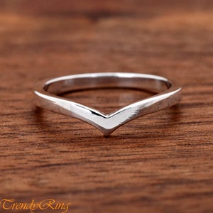 May include: A silver-colored chevron ring. The ring has a V-shaped design and is set against a wooden background. The ring is a simple, elegant piece of jewelry.