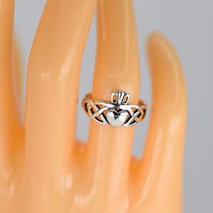 May include: A silver Claddagh ring featuring a heart, crown, and hands design. The ring is on a finger, showcasing the intricate Celtic knotwork around the heart. The ring's design is a symbol of love, loyalty, and friendship.