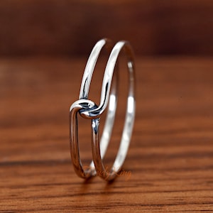 May include: A silver ring with a knot design. The ring features two bands that are joined together with a knot in the center. The ring is made of a shiny, polished metal and is set against a wooden background.