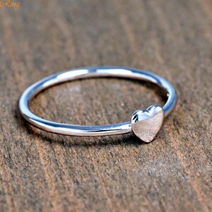 May include: A thin, polished silver-tone ring featuring a small, smooth heart-shaped charm attached to the band. The metal surface is reflective and metallic, showcasing a simple, continuous circular design with the text TrendyRing in the corner.