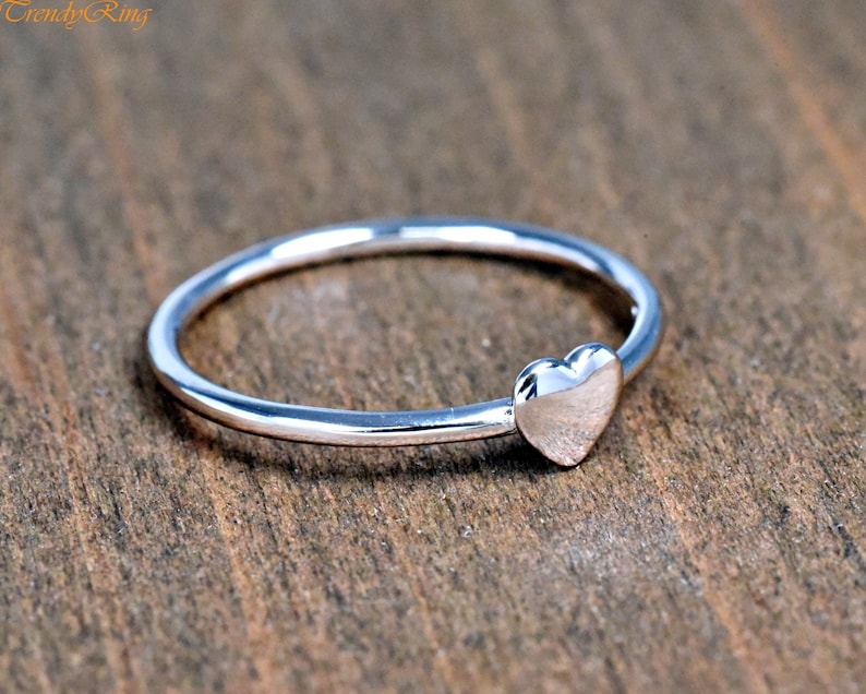 May include: A thin, polished silver-tone ring featuring a small, smooth heart-shaped charm attached to the band. The metal surface is reflective and metallic, showcasing a simple, continuous circular design with the text TrendyRing in the corner.