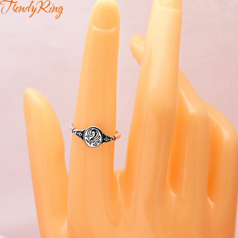 May include: A silver-toned ring featuring an oval-shaped centrepiece with a black engraved swirling pattern. The band is thin and metallic, displaying intricate detailing on the sides that connects to the central oval element.