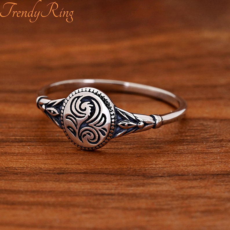 May include: A silver-toned ring featuring an oval-shaped centrepiece with an engraved swirling floral pattern. The band is thin and polished, with decorative leaf-like engravings on the sides leading to the central oval element. The text Trendy Ring is visible in the upper left corner.