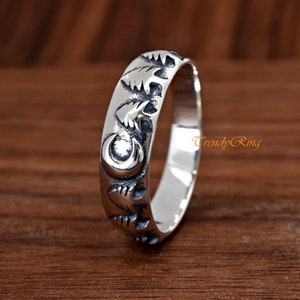 May include: A silver band ring with a design of three pine trees and a crescent moon. The ring is on a wooden surface.