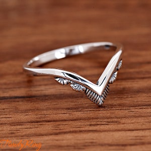 May include: A silver-colored ring with a V-shaped design. The ring has a decorative pattern along the V-shape and is set against a wooden surface. The ring is a piece of jewelry.