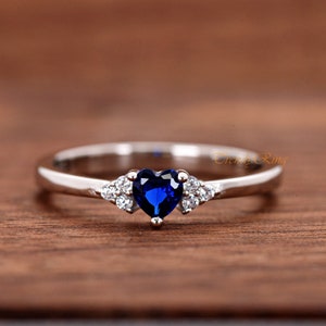May include: A silver ring with a blue heart-shaped gemstone and smaller clear gemstones on the band. "Trendy Ring" is written in the background.