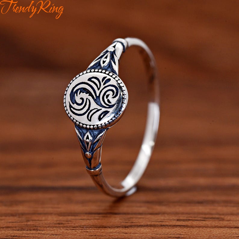 May include: A silver-toned ring featuring a circular centrepiece with a dark, recessed swirling pattern. The band is adorned with intricate, oxidised metal detailing that extends from the central disc, creating a textured, metallic appearance.