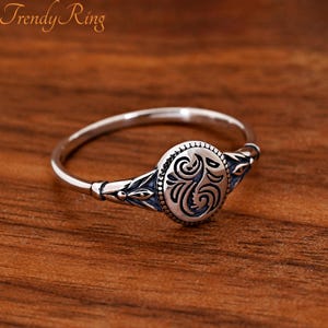 May include: A silver ring featuring a central oval plaque with an engraved swirling pattern and beaded border. The band displays decorative leaf-like motifs on both sides, with oxidised recessed areas that highlight the intricate metalwork details.