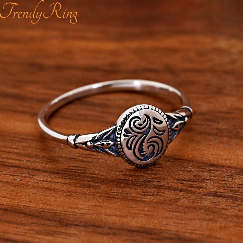 May include: A silver ring featuring a central oval plaque with an engraved swirling pattern and beaded border. The band displays decorative leaf-like motifs on both sides, with oxidised recessed areas that highlight the intricate metalwork details.