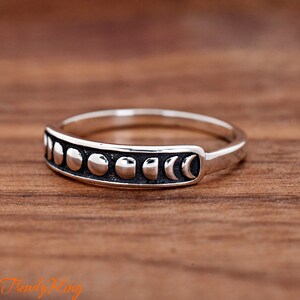 May include: A silver-toned ring featuring a band with a series of raised, polished moon phase symbols set against a dark, recessed background. The design displays the lunar cycle from a full moon to a crescent moon across the front of the band.