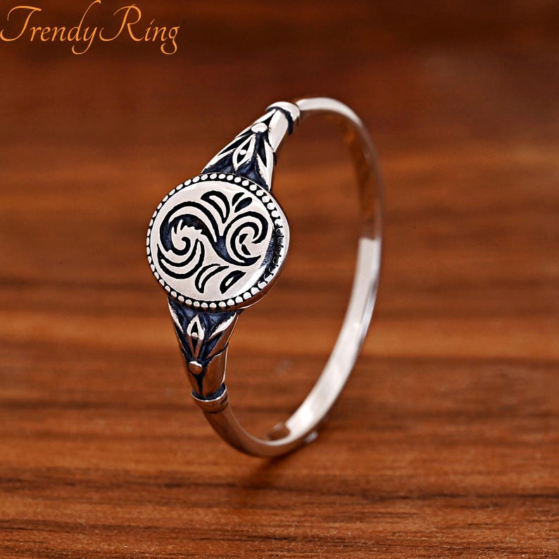 May include: A silver-toned ring featuring a circular centrepiece with a black engraved swirl pattern and a beaded border. The band displays decorative floral-inspired engravings on the shoulders, and the text Trendy Ring is visible.