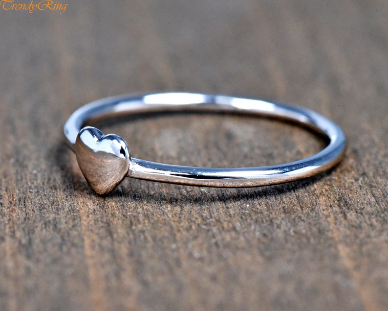 May include: A polished silver-toned ring featuring a small, smooth heart-shaped charm attached to a thin, circular band. The text "TrendyRing" is visible in the upper left corner of the image, presented in a simple, orange-colored font.