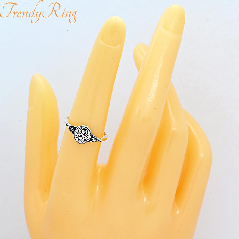 May include: A silver-coloured ring featuring a small, oval-shaped face with an intricate black engraved swirl pattern. The band is thin and metallic, and the text Trendy Ring is visible in the upper left corner of the image.