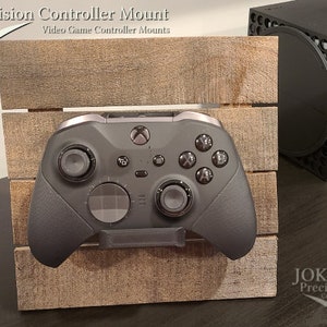 May include: A black Xbox controller with a gray faceplate is mounted on a wooden stand. The stand is made of reclaimed wood and has a rustic look. The controller is mounted on the stand using a 3D printed mount. The stand is labeled "Joker Precision Co."