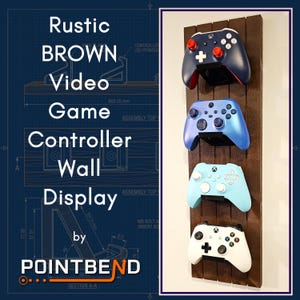 May include: A vertical dark brown wooden wall mount display rack featuring four black brackets holding four different video game controllers. The controllers are colored dark blue with red accents, metallic blue, light blue, and white with black accents.