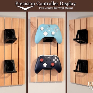 May include: Two black wall mounts for displaying video game controllers. The mounts are attached to a wooden wall. Two controllers are displayed on the mounts, one blue and one dark blue.