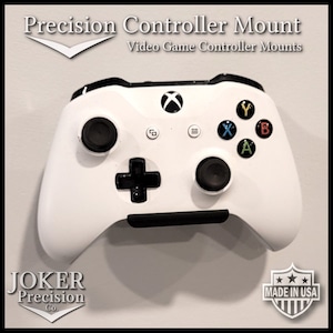 May include: White Xbox controller mounted on a wall with a black mount. The controller has black buttons and a black D-pad. The text "Precision Controller Mount" and "Video Game Controller Mounts" is at the top of the image. The text "JOKER Precision Co." is at the bottom left of the image. The text "MADE IN USA" is at the bottom right of the image.