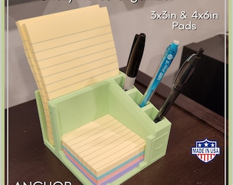 May include: A green plastic sticky note organizer with three slots for pens and a compartment for sticky note pads. The organizer is holding a stack of 3x3 inch and 4x6 inch sticky note pads. The organizer is on a wooden surface. The text "Anchor Precision" and "Made in USA" are visible in the image.