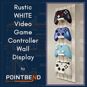 May include: A vertical white wooden wall display rack featuring four individual white mounts for video game controllers. The display holds four controllers in black, metallic blue, light blue, and white colors. Text reads: Rustic WHITE Video Game Controller Wall Display by POINTBEND.