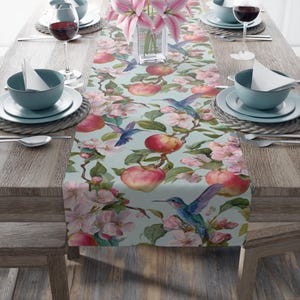 May include: A light blue table runner with a watercolor design of red apples, pink blossoms, and hummingbirds. The runner is set on a wooden table with place settings, wine glasses, and a vase of pink lilies.