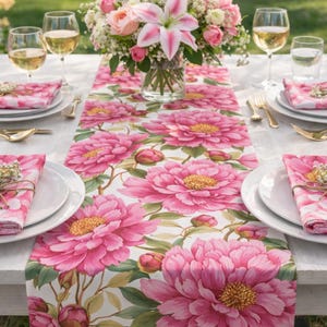 May include: A floral table setting featuring a white table runner with large pink peony blooms and green leaves. White plates, napkins, and gold silverware are arranged around the runner. A vase of flowers and wine glasses complete the scene.
