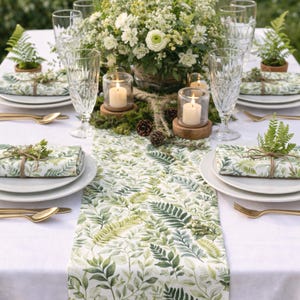 May include: A beautifully set table with a white tablecloth, featuring a green leaf-patterned runner and matching wrapped gifts. Gold cutlery, clear glasses, and a floral centerpiece complete the elegant outdoor dining scene. Candles in glass holders add a warm glow.