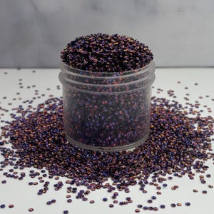 May include: A clear plastic jar filled with iridescent, dark purple sequins. The sequins are scattered around the jar on a white surface. The sequins have a metallic sheen with hints of gold and blue.
