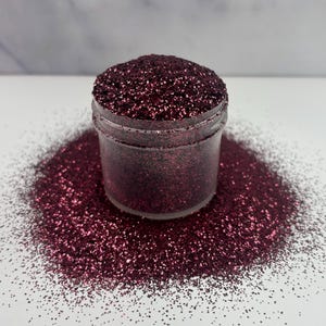 May include: A glass jar filled with fine burgundy glitter. The glitter is scattered around the jar on a white surface.