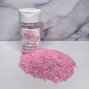 May include: A bottle of pink glitter labelled "Cactus Blossom" sits beside a pile of the same glitter. The glitter is a mix of pink and iridescent particles. The bottle has a white lid and a label with a logo and product information.
